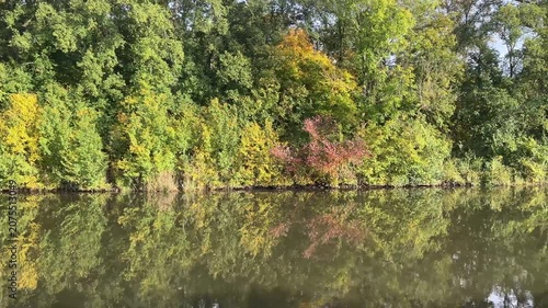 colorful river landscape seen from a boat in autumn, trees reflected in the river, piled stones on the bank, a bench next to the river, yellow-green leaves on the trees, beautiful green reflection