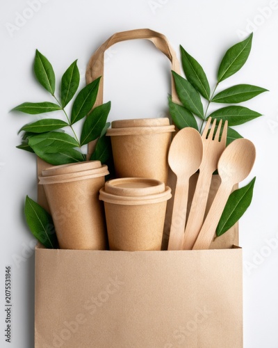 Eco paper cups wooden cutlery and green leaves in kraft bag on white background