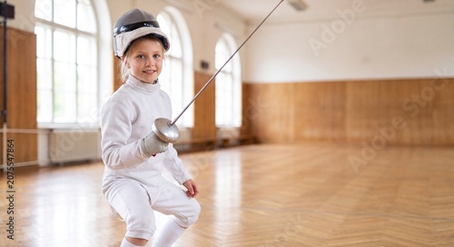 A focused young girl wearing a fencing uniform and mask stands in the en garde position with her foil in a sports hall.