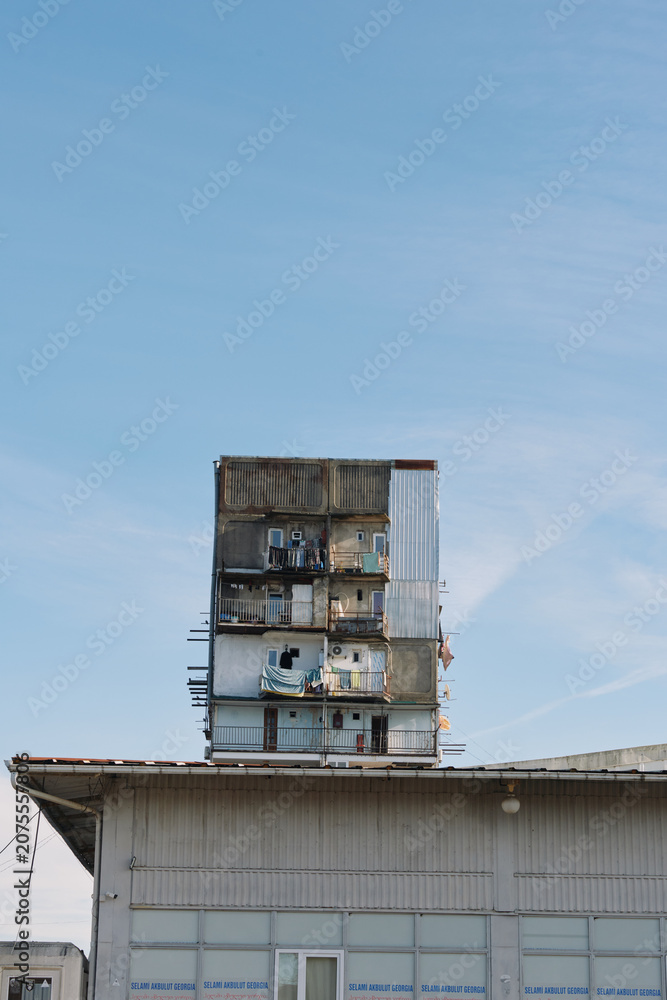 Fototapeta premium Tall stacked apartment tower atop a low industrial building under a clear blue sky, with small figures on balconies, urban housing structure and exposed living spaces.