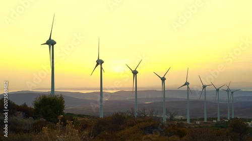 A row of wind turbines stands on a hillside as the sun sets behind them, generating clean energy. The turbines' white blades rotate in the breeze, surrounded by sparse vegetation.