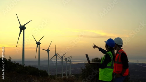 Two workers in safety gear, one wearing a blue hard hat and high-visibility vest, the other in a white hard hat and orange vest, examine wind turbines on a hillside at dusk.