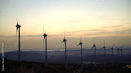 A row of wind turbines stands on a hillside as the sun sets behind them, generating clean energy. The turbines' white blades rotate in the breeze, surrounded by sparse vegetation.