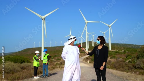 A man in traditional Middle Eastern attire and a woman in business casual discuss wind turbine operations with two technicians in hard hats and safety vests on a rural road