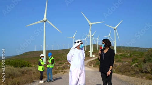 A man in traditional Middle Eastern attire and a woman in business casual discuss wind turbine operations with two technicians in hard hats and safety vests on a rural road