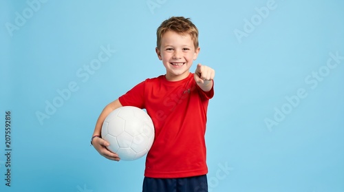 Happy young boy in red t-shirt holding a volleyball and pointing at the camera on a blue background.
