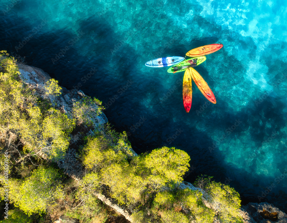 Fototapeta premium Aerial drone view of colorful kayaks in crystal clear turquoise sea near rocky coast and green trees. Summer outdoor adventure in Mallorca, Balearic Islands, Spain. Top down view of canoes. Travel