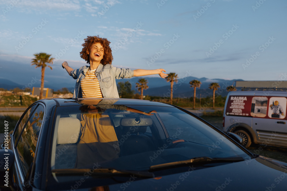 Fototapeta premium Joyful young woman leans from a car sunroof with arms wide in a sunlit coastal lot, capturing carefree travel vibes.