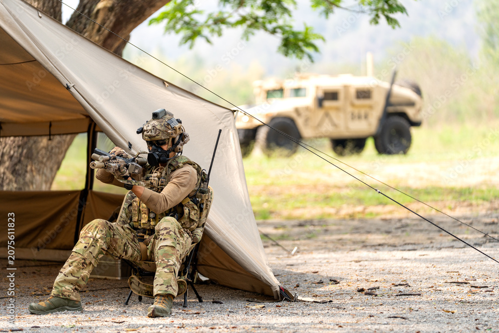 Fototapeta premium One army man or soldier sit on chair and check his weapon in front of military tent with High Mobility Multipurpose Wheeled Vehicle in the background.