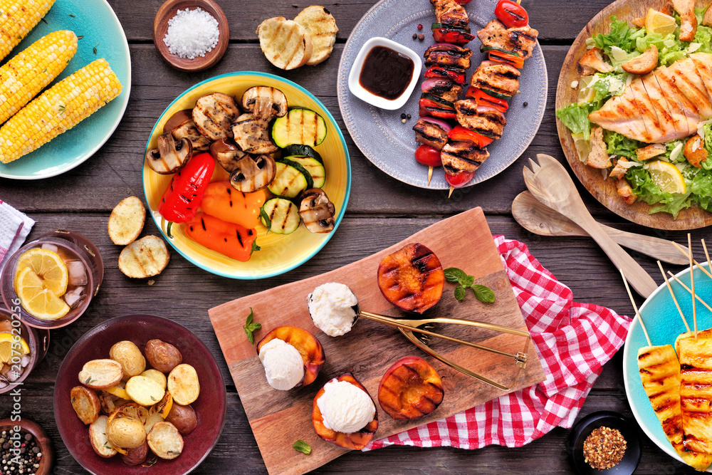 Fototapeta premium Summer BBQ food table scene over a dark wood background. Grilled meat skewers, corn, potatoes, vegetables, pineapple, peaches and ice cream, salad and iced tea. Top view.