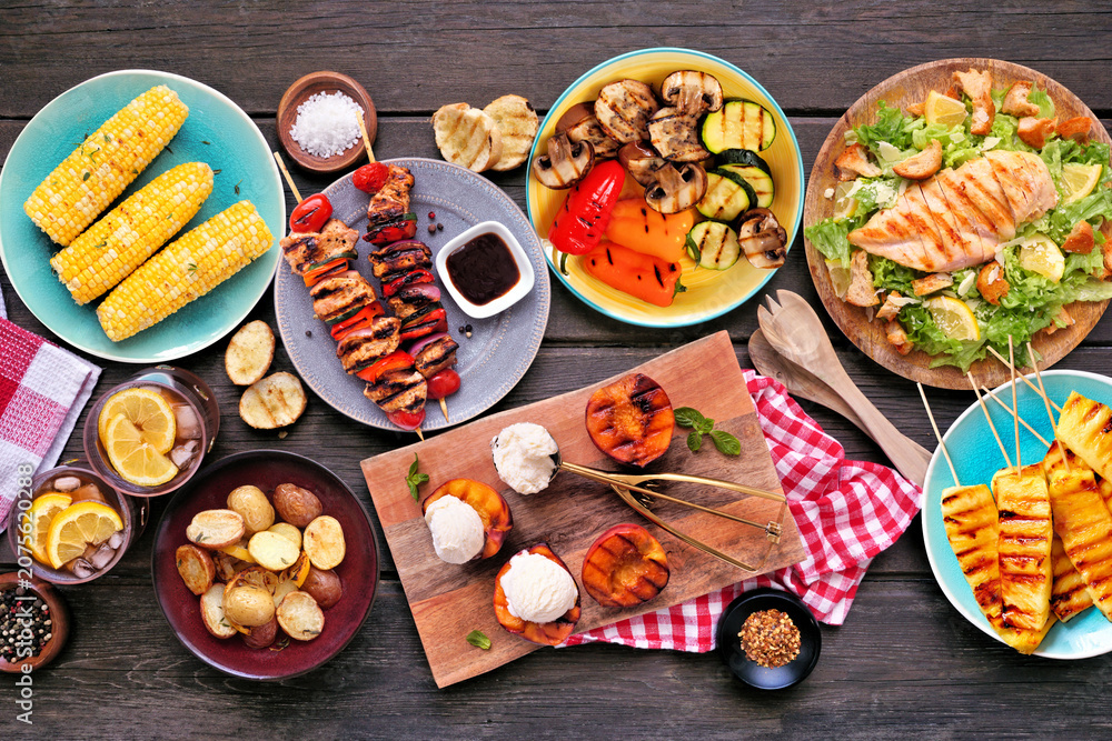 Obraz premium Summer BBQ food table scene over a dark wood background. Grilled meat skewers, corn, potatoes, vegetables, pineapple, peaches and ice cream, salad and iced tea. Overhead view.
