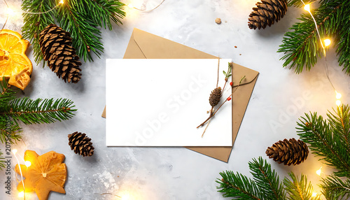 blank white christmas card mockup with craft envelope surrounded by evergreen pine branches, pinecones and warm fairy lights on rustic surface