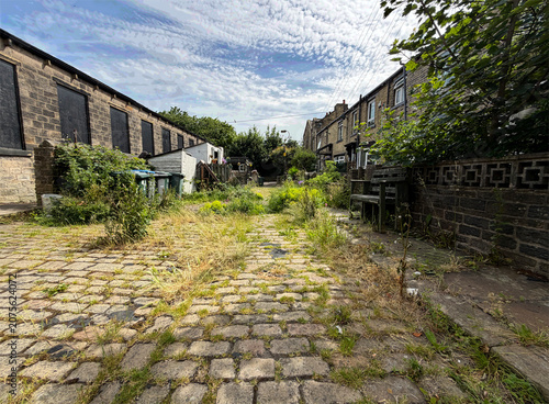 A Victorian cobblestone street winds between weathered stone buildings, their edges softened by creeping weeds, as scattered clouds drift lazily above the High Street in Wibsey, Yorkshire, UK.