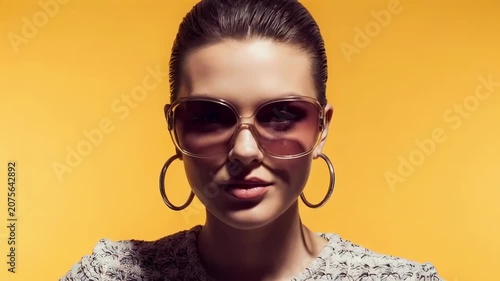 Fashion portrait of a stylish woman in oversized sunglasses and hoop earrings against a vibrant yellow background, posing and turning