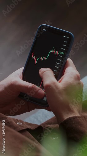 Close-up of a trader hands holding a smartphone displaying a cryptocurrency candlestick chart. Investor tracks stock market or crypto data and exchange rates on a mobile trading app. Vertical video