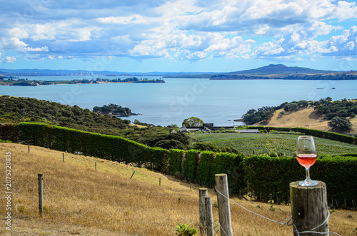 A view towards Auckland New Zealand and Rangitoto volcano from Waiheke Island, New Zealand. A glass of wine from a local winery rests on a fence pole, completing the view