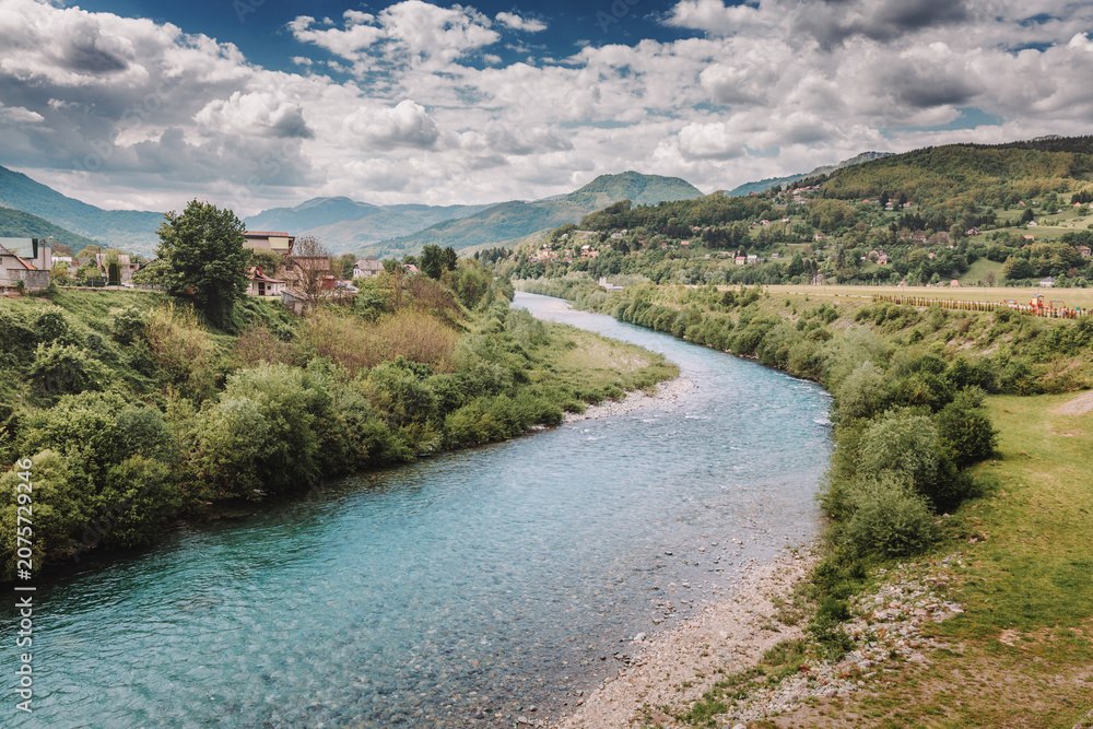 Obraz premium Blue Tara river winding through a lush valley, with village houses nestled on hillsides and a cloudy sky above the mountains in Montenegro, Mojkovac.