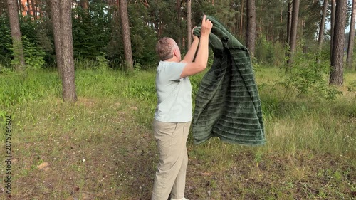 A mature woman shakes out a dirty picnic blanket in a summer forest, outdoor recreation and a picnic in nature