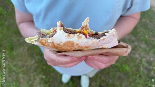 A woman holds a half-eaten grilled meat wrap outdoors, a close-up of street-food shawarma in a park