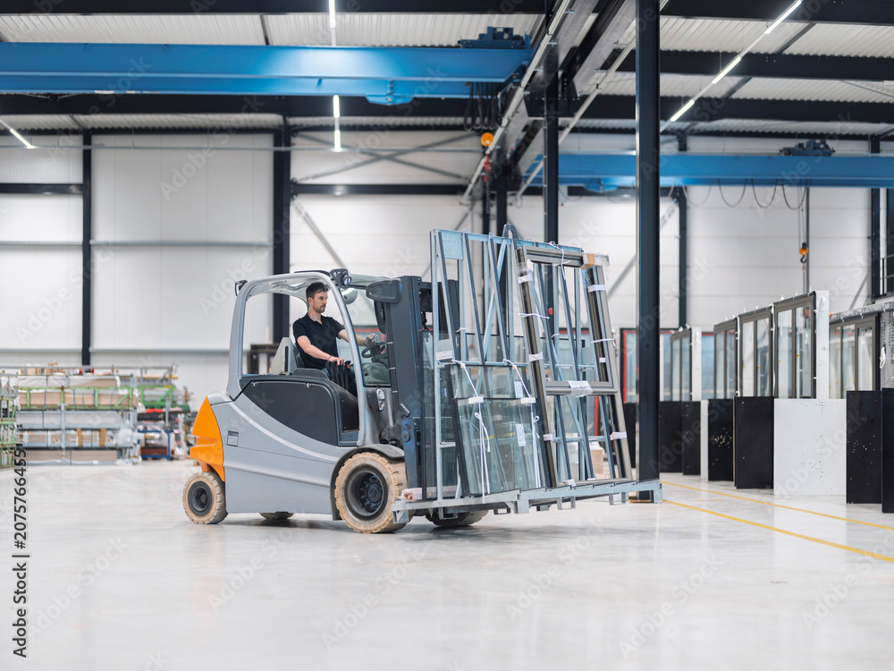Fototapeta premium Forklift driver moves an A-frame rack of insulated glass through a glazing warehouse, the cab seen in near profile.