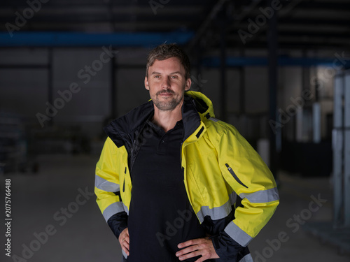 Worker stands with hands on hips in a hi-vis jacket, a slight smile and assertive stance in a dim warehouse.