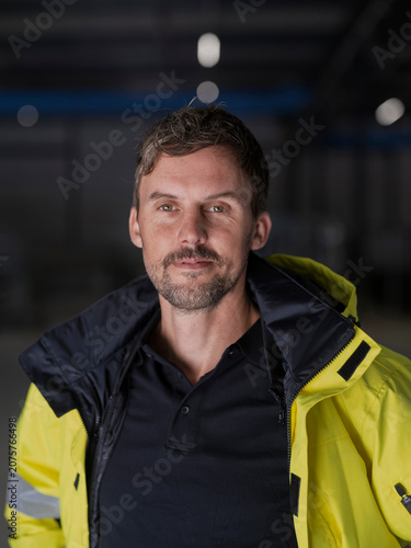 Tight head-and-shoulders portrait of a bearded worker in a hi-vis jacket, soft light bokeh behind in a dark hall.