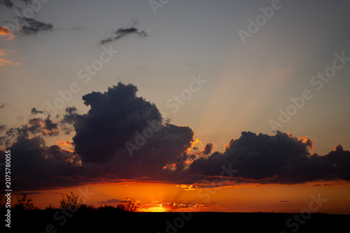 Photography Bright sun setting behind the horizon with dark cumulus clouds and visible sunbeams or crepuscular rays spreading across the evening sky