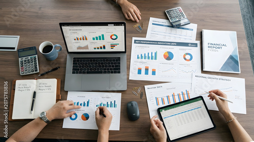 Overhead view of a business team collaborating on financial data analysis with laptops, tablets, and paper documents displaying charts and graphs.