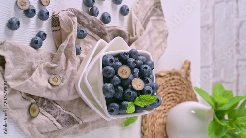 A white square bowl filled with plump blueberries sits on a rustic linen cloth, featuring one berry sliced in half to show its creamy interior under bright natural light.