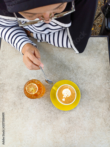 Hijab woman with coffee latte cup and iced lemon tea glass on outdoor cafe table. Female model holding spoon over beverages, high angle top view.