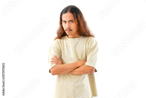 A studio portrait of a young man with long hair and a mustache, wearing a beige t-shirt, standing with his arms crossed and looking at the camera, isolated on white background.