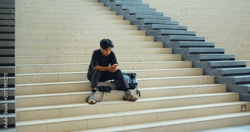 Young man seated on stair steps, looking at smartphone, backpack and camera beside him in modern interior, calm urban lifestyle moment with wide symmetrical composition and soft natural light