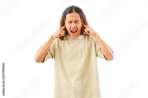 A young man with long hair wearing a beige t-shirt, covering his ears with his fingers and screaming with his mouth wide open, isolated on white background.