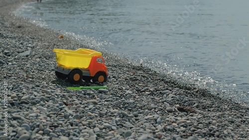 Bright yellow plastic dump truck green shovel rest on gray pebble beach beside gentle sea waves. Soft natural daylight highlights textures