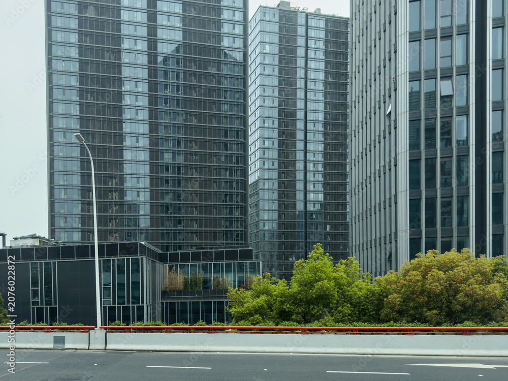 Obraz premium Glass towers behind roadside trees and traffic barrier in Shanghai, China, spring day