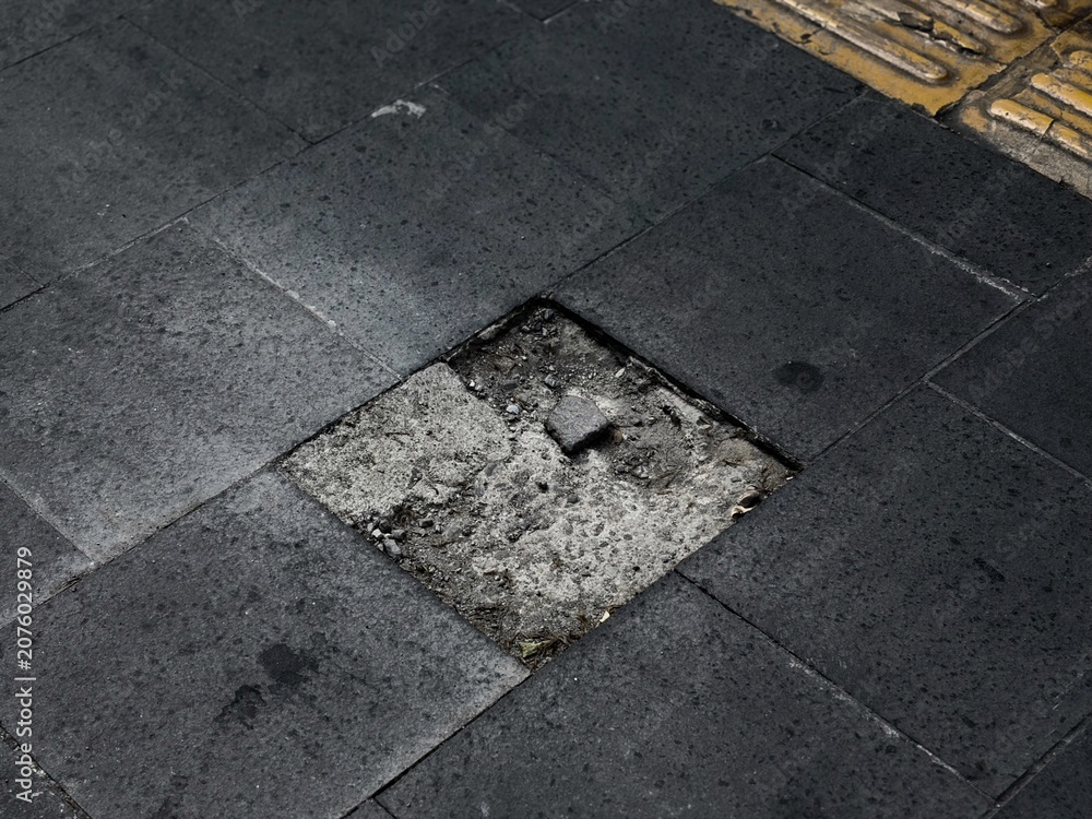 Fototapeta premium Missing black sidewalk tile exposing broken concrete and debris, a tripping hazard on urban pavement
