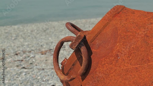 Weathered big buoy with corroded metal rings on gray pebble shore. Soft natural daylight highlights texture. Calm sea in background. Evokes abandonment, decay, coastal solitude
