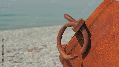 Close up of weathered rusted buoy with metal ring on pebble shore. Soft natural daylight highlights texture. Calm sea in background. Evokes abandonment, coastal decay, nautical themes