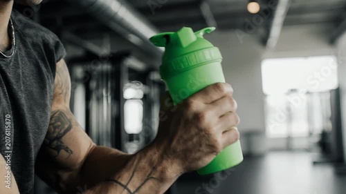 Male fitness enthusiast with tattoos shakes a green shaker bottle in a gym, showcasing a focus on hydration and workout preparation in a modern fitness environment