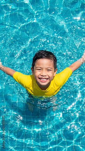 Asian boy with a big smile swims in a blue pool on a sunny day, top view. Fun summer vacation and travel concept