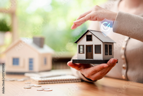 Home insurance and property protection concept. Woman hands holding miniature house model with secure shield checkmark icon.