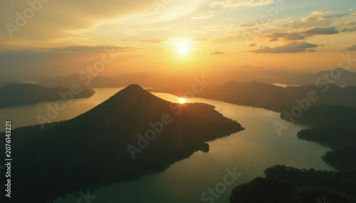 Aerial view of a mountain peak surrounded by river water during sunset. Golden hour landscape with lake and hills. Scenic nature background for travel and serenity environment concept.