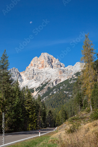 Mountain road leading to the Dolomite peaks under deep blue sky with daytime moon, Italy