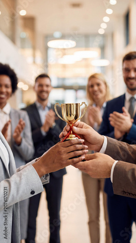 Close-up of a Black woman and Caucasian man holding a golden trophy cup. A diverse group of blurred business colleagues in suits applaud and celebrate their success in a bright office hall.