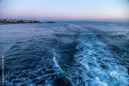 Foam wake trace on blue sea water behind a ferry boat boat in Istanbul. Ship track wave pattern in sunset twilight, maritime transport, evening travel background, April, Turkey.