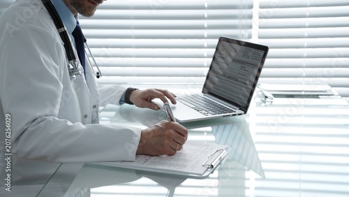 Photography Doctor sitting at a desk, typing on a laptop with a stethoscope around their nec