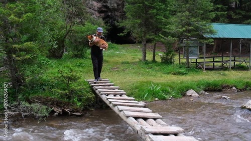 Man with backpack carefully helping pembroke welsh corgi dog cross narrow wooden footbridge over mountain river. Concept for outdoor adventure travel, active lifestyle, pet care and companionship.