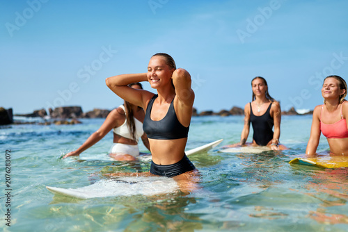 Group of young women sit on surfboards in clear ocean water, smiling and relaxing after surfing workout. Fit female friends enjoy sunny beach day, active lifestyle, blue sky.