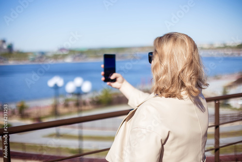 Woman with light blonde hair takes a picture with her smartphone, overlooking a river from a balcony on a bright sunny day