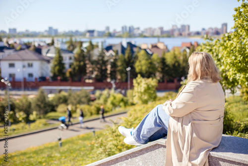 Woman with blonde hair sits on a stone ledge, gazing at a city and river view, with trees and houses visible in the background